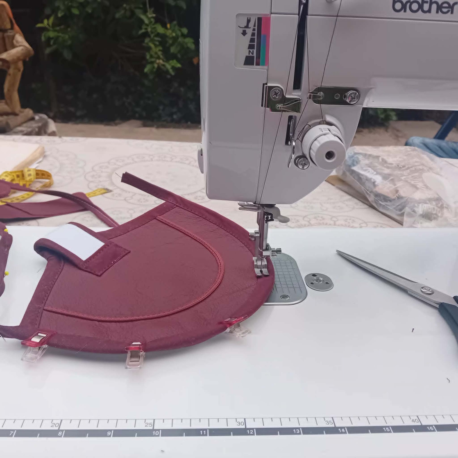 Een Brother-naaimachine waarmee een handgemaakt bordeauxrood lederen kippenzadel van Mammientje Tien nauwkeurig wordt gestikt. Op de werktafel liggen naai-benodigdheden zoals stofklemmen, spelden en een stofschaar.